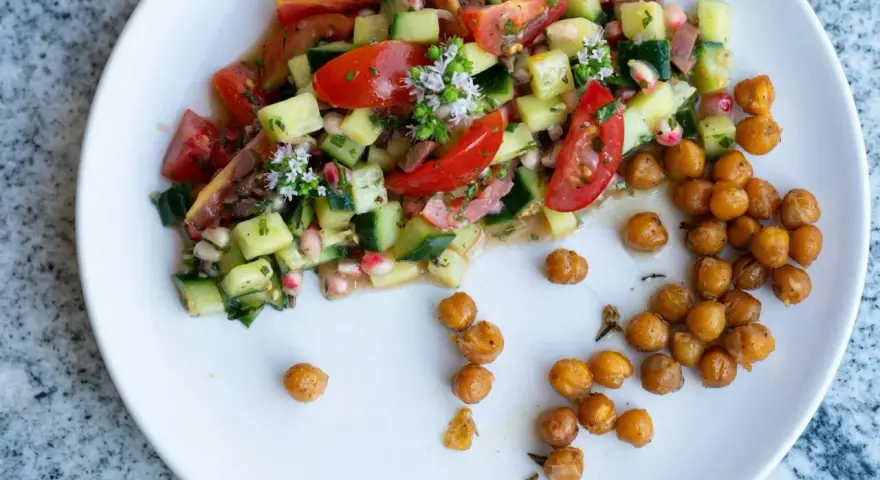 Salade colorée de tomates, concombres et pois chiches, garnie de petites fleurs blanches. Des pois chiches rôtis sont disposés à côté.