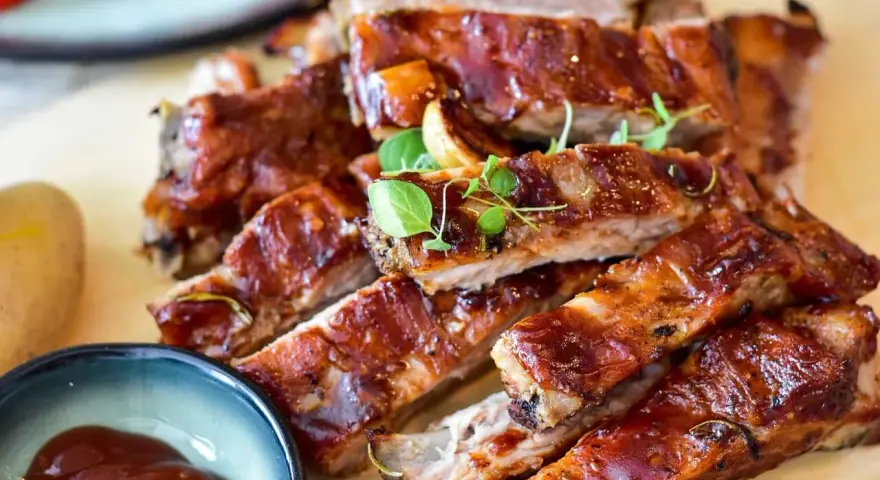 Délicieux ribs au four, nappés d'une sauce barbecue brillante, servis avec une petite coupelle de sauce et des herbes fraîches.
