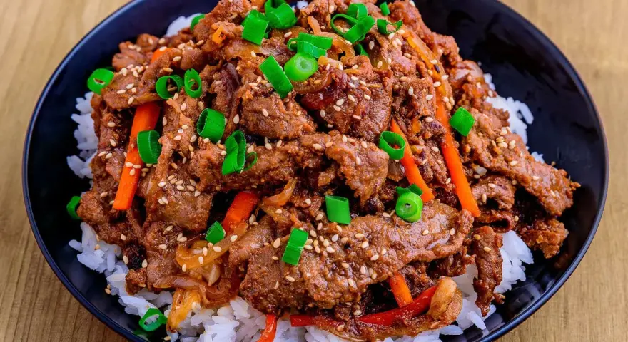 Un bol de riz blanc garni de boeuf bulgogi mariné, de julienne de carottes et d'oignons verts ciselés, parsemé de graines de sésame.