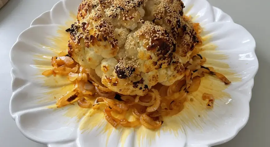 Un chou-fleur entier, rôti au four, est servi sur une assiette blanche ornée d'oignons caramélisés. Le chou-fleur est saupoudré de graines de sésame et d'épices.