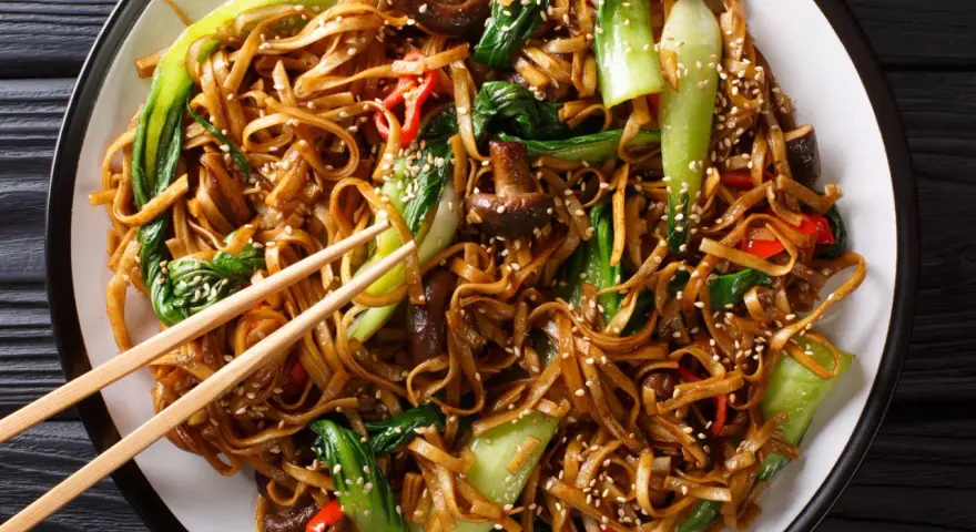 Un plat de chow mein appétissant avec des nouilles, des légumes verts croquants, des champignons et des poivrons rouges, saupoudré de graines de sésame.