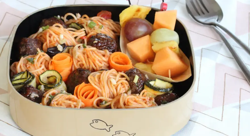 Bento box avec spaghetti et boulettes, garni de courgettes et carottes roulées. Une brochette de fruits complète cette idée repas à emporter au travail.