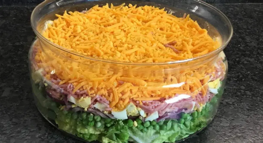 Salade à étage colorée dans un bol en verre : laitue, petits pois, oignons rouges, jambon, œufs durs et fromage râpé.
