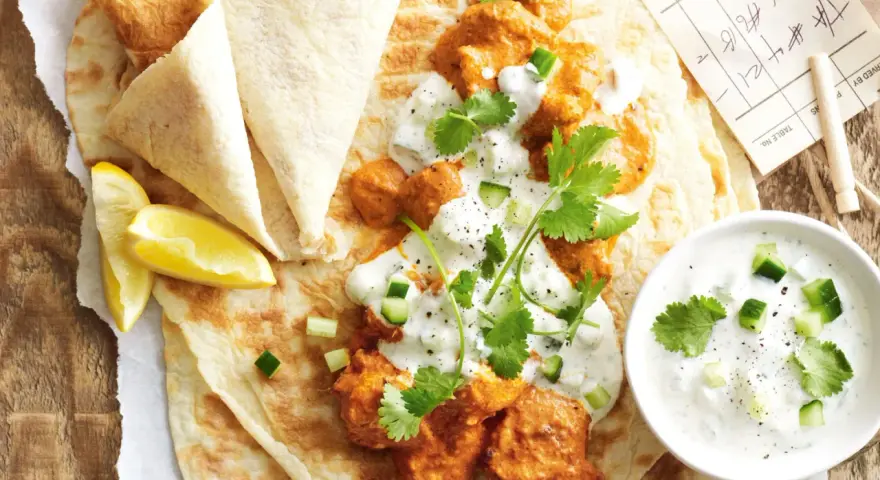Un plat de butter chicken crémeux servi sur du pain naan avec du raïta et des quartiers de citron.