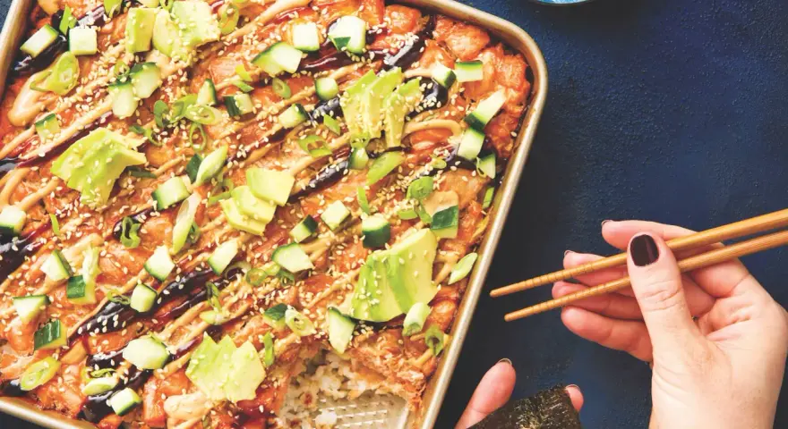 Un délicieux sushi bake dans un plat doré, garni d'avocat, concombre, oignons verts et graines de sésame. Une main tient des baguettes pour servir.