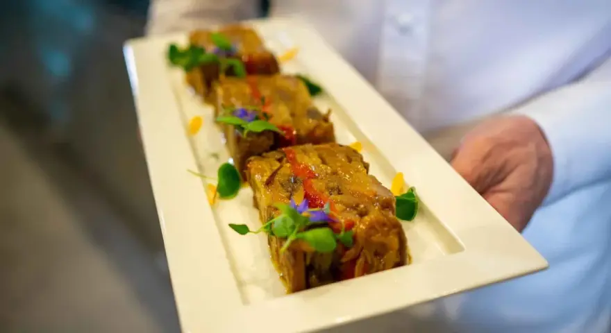 Trois tranches de terrine de légumes, garnies de fleurs comestibles et de verdure, présentées sur une assiette blanche par un serveur.