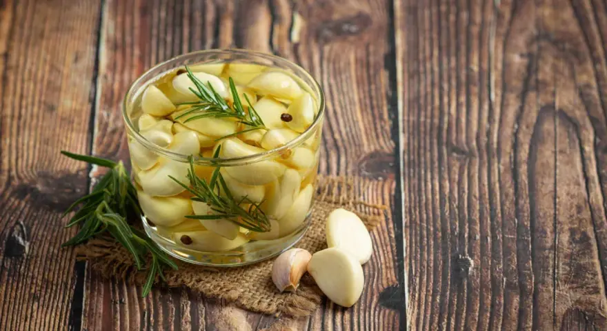 Un bocal de **ail confit** avec des brins de romarin et quelques grains de poivre, posé sur une toile de jute, sur un fond de bois rustique.