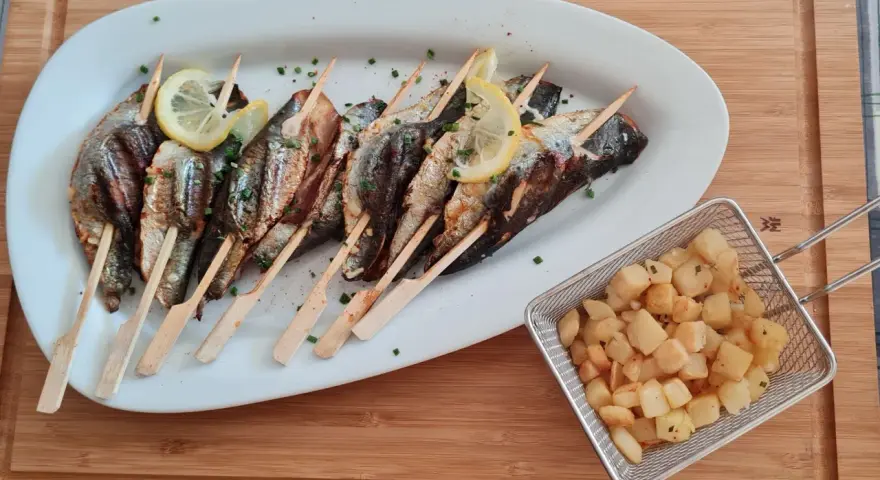 Sardines grillées sur brochettes, garnies de citron et persil, accompagnées de pommes de terre sautées dans un panier métallique.