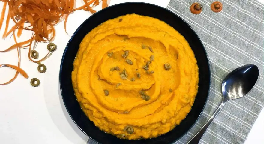 Un bol de purée de carotte onctueuse, garnie de graines, à côté d'une cuillère et de carottes râpées.