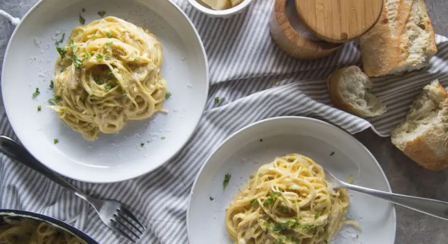 Deux assiettes de pâtes crémeuses à l'ail, garnies de persil frais. Un pain croustillant et du fromage râpé complètent cette recette de pâtes à l'ail et crème.