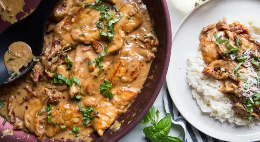 Poulet en sauce tomate séchée crème, garni de basilic frais, servi dans une poêle et sur une assiette de riz.