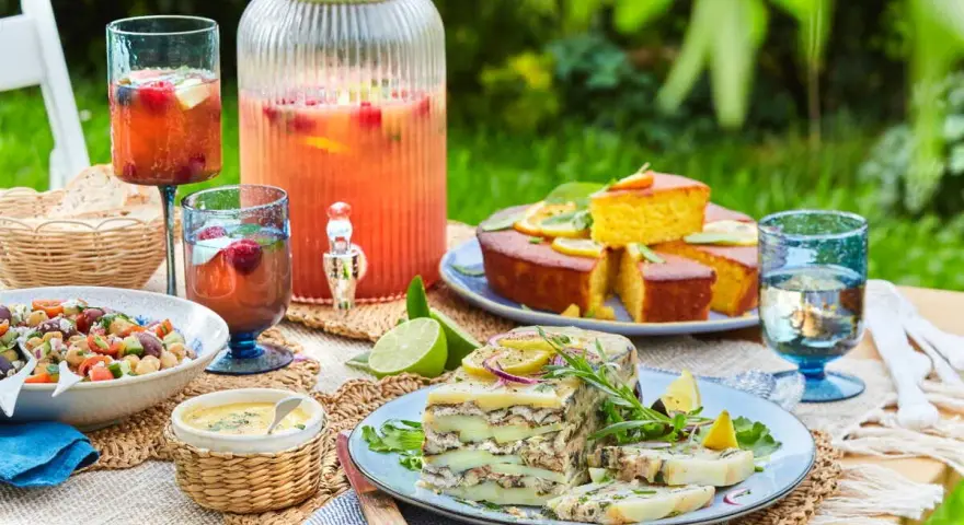 Un délicieux repas d'été entre amis : salade, terrine, gâteau, et une grande carafe de boisson fruitée, le tout sur une table dressée en extérieur.