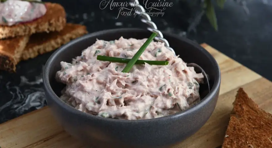 Un bol de rillettes de thon crémeuses, garni de ciboule, prêt à être tartiné sur des toasts grillés.
