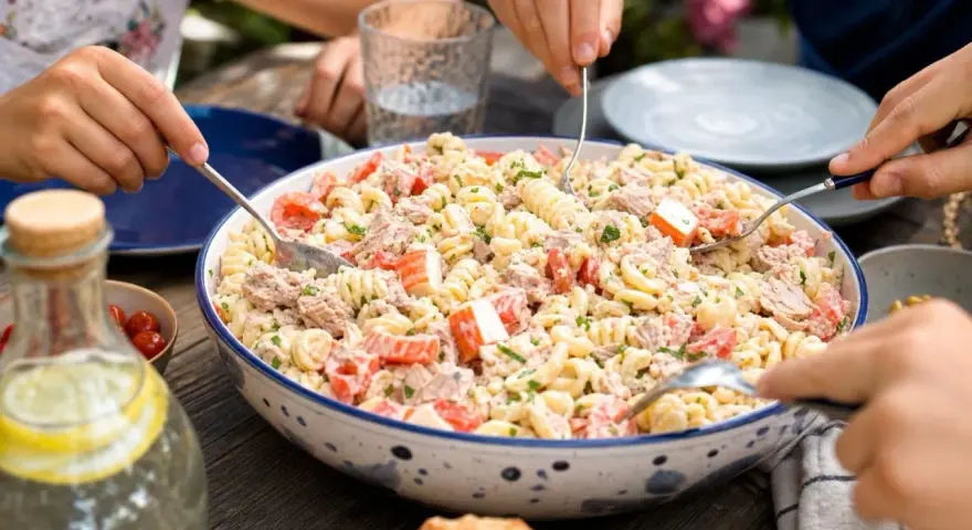 Un grand plat de salade de pâtes au surimi, tomates et thon, servi à l'extérieur. Des mains servent la salade.