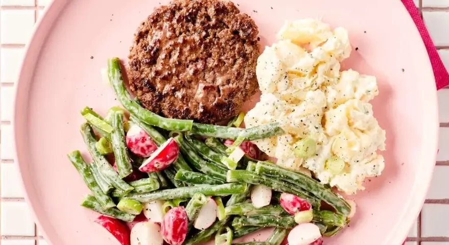 Un hamburger savoureux avec une salade de haricots verts et radis, et une salade de pommes de terre crémeuse sur une assiette rose.