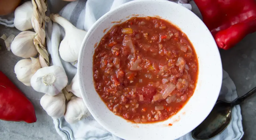 Un bol de salsa facile, avec des tomates, des oignons et des poivrons rouges. Des gousses d'ail et un oignon sont disposés autour.