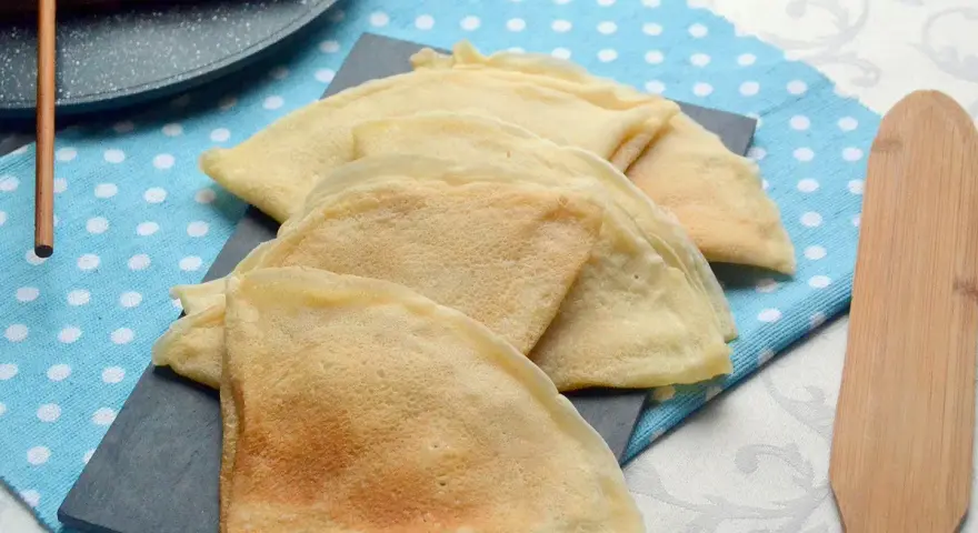 Pile de crêpes dorées, prêtes à être garnies. La pâte à crêpe vegan est légère et aérienne. Un ustensile en bois est à côté.