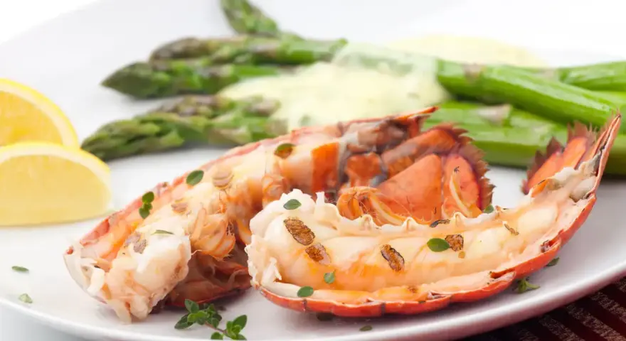 Queue de homard grillée, servie avec des asperges et une sauce homard crémeuse, garnie de quartiers de citron et de thym frais.