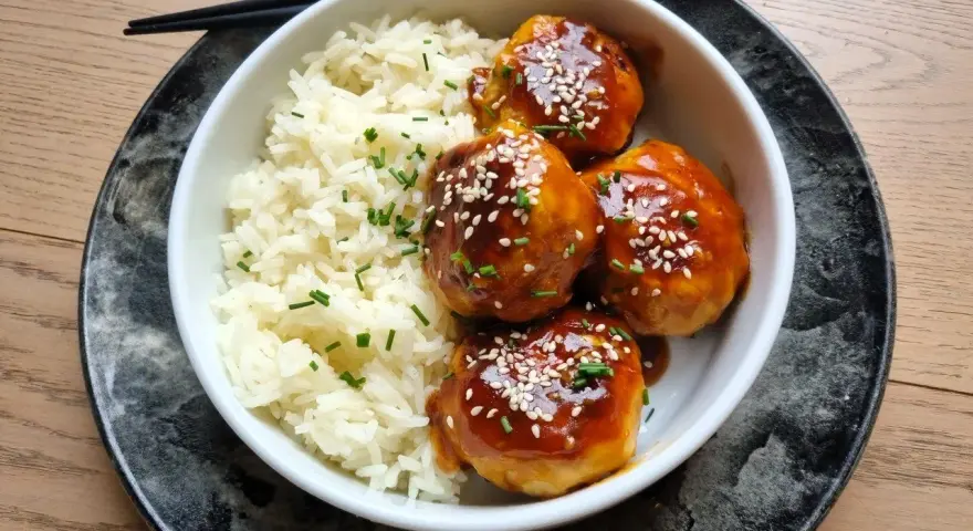 Délicieuses boulettes de poulet asiatique nappées d'une sauce sucrée et salée, servies avec du riz blanc et parsemées de graines de sésame.