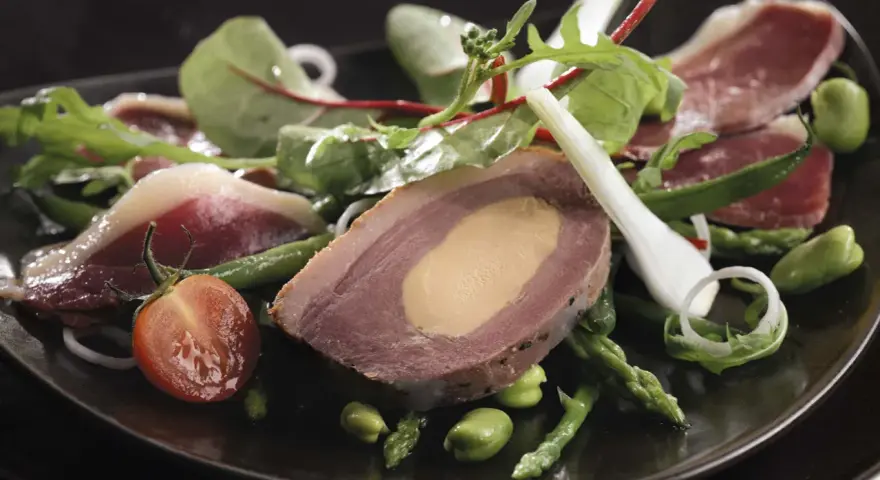 Une délicieuse salade landaise avec du foie gras, du magret de canard, des asperges et des tomates cerises sur une assiette noire.