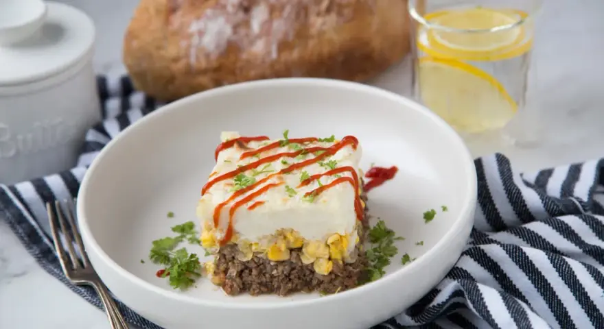 Un délicieux pâté chinois servi dans une assiette blanche, garni de persil et de ketchup. Un verre d'eau citronnée et du pain complètent ce plat réconfortant.