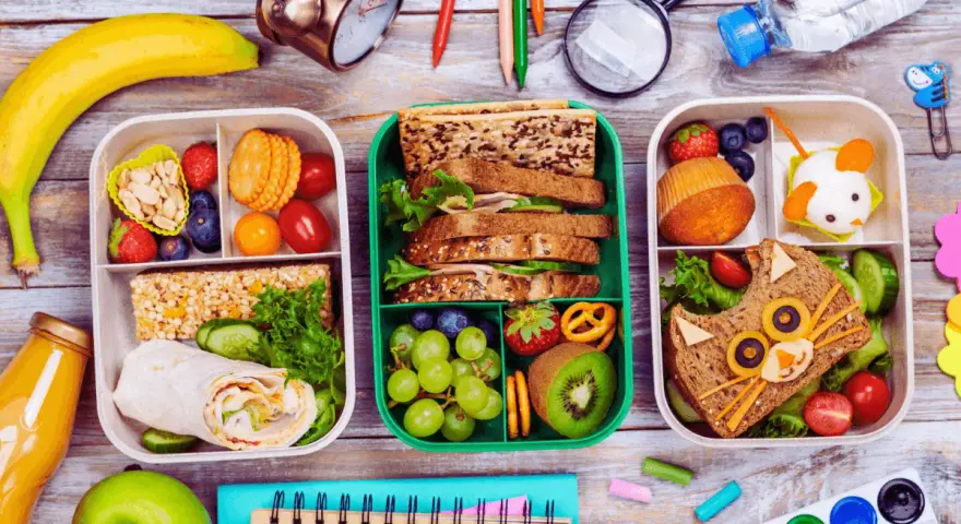 Trois boîtes repas à emporter faites maison, remplies de fruits, sandwichs créatifs et barres de céréales. Parfait pour un pique-nique ou un déjeuner sur le pouce.