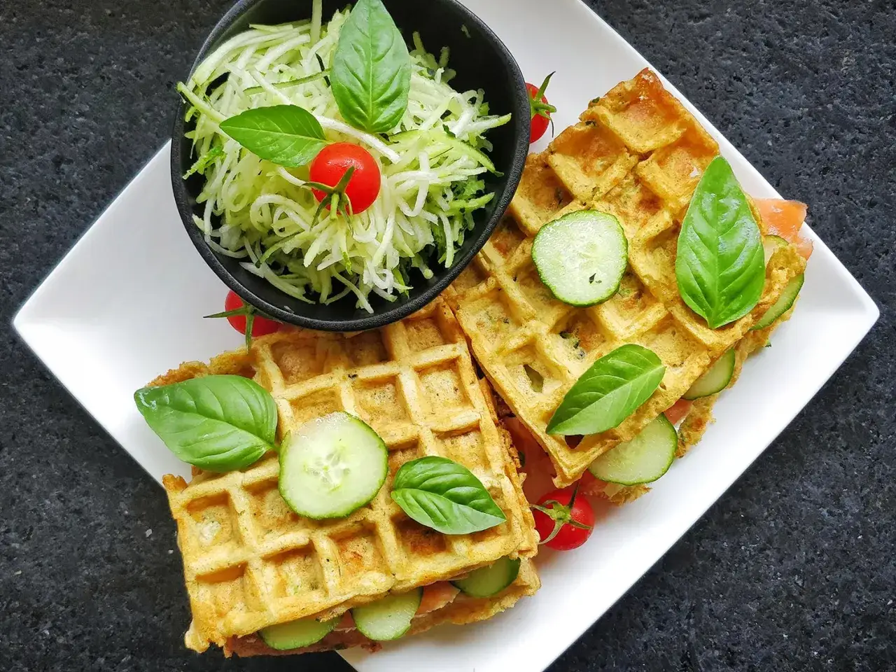 Deux gaufres salées garnies de concombre et de basilic, accompagnées d'une salade de courgettes râpées et de tomates cerises.