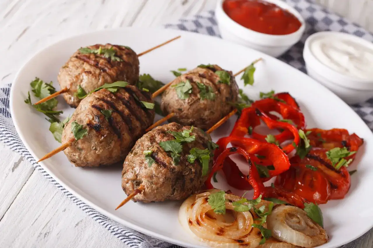 Brochettes de kefta grillées, garnies de coriandre fraîche, accompagnées de poivrons rouges rôtis et d'oignons caramélisés. Une recette parfaite pour un repas savoureux.