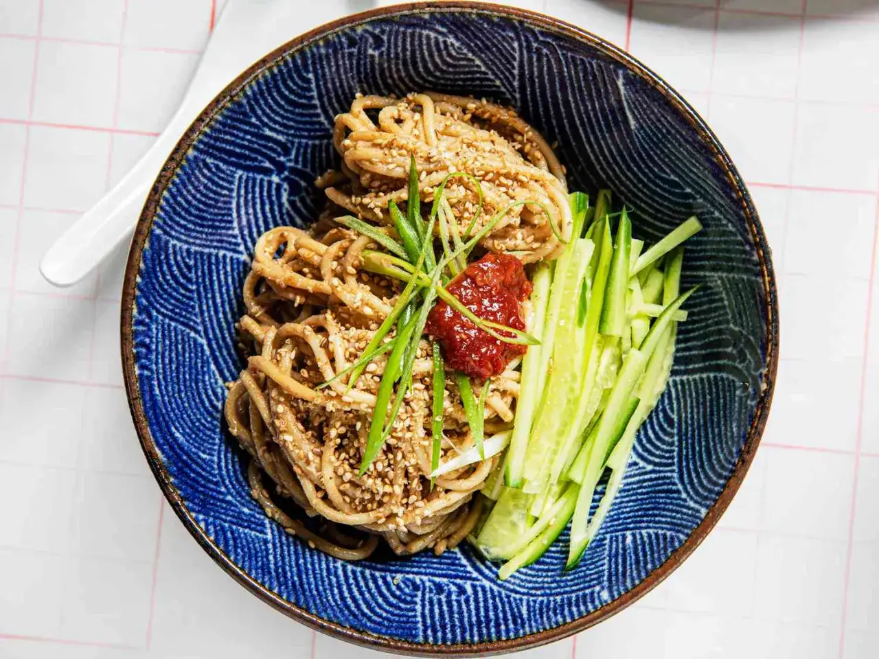 Salade asiatique de nouilles avec concombre, oignon vert et p&acirc;te de piment, garnie de graines de s&eacute;same dans un bol bleu.
