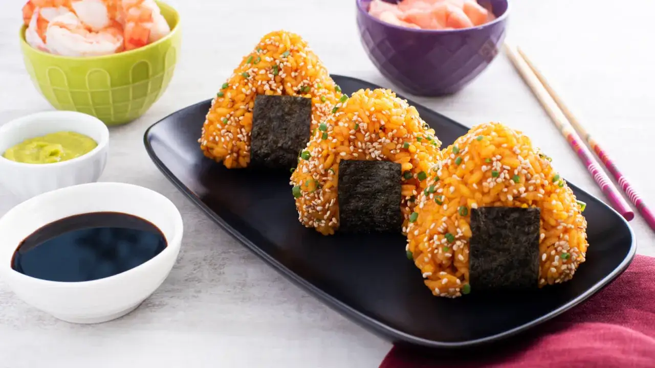 Trois onigiri crevette, enrob&eacute;s de s&eacute;same et d'oignons verts, sur une assiette noire avec des accompagnements.