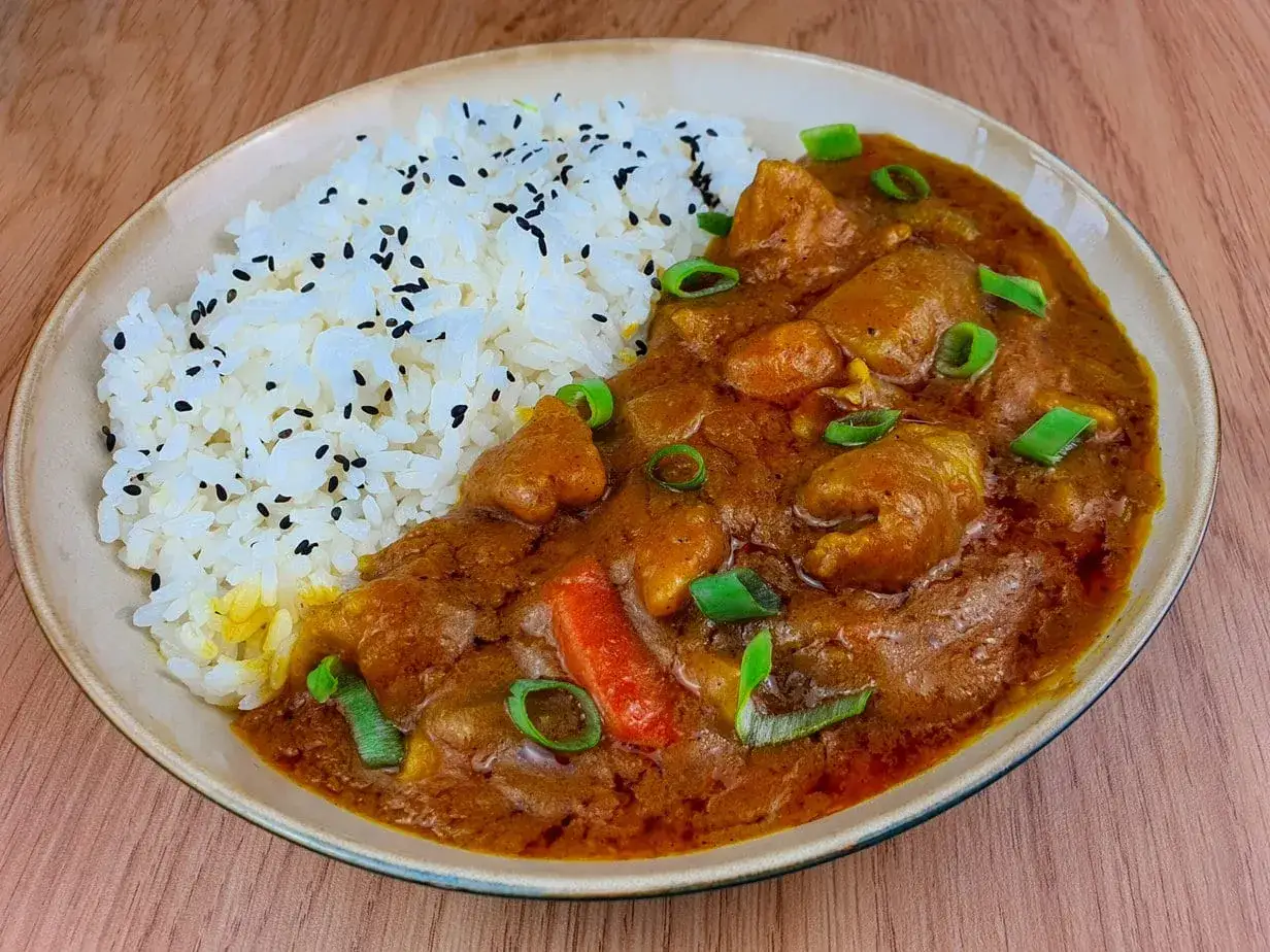 Un plat de riz blanc parsemé de graines de sésame noires, servi avec un savoureux curry japonais aux morceaux de poulet et légumes, garni de ciboule.
