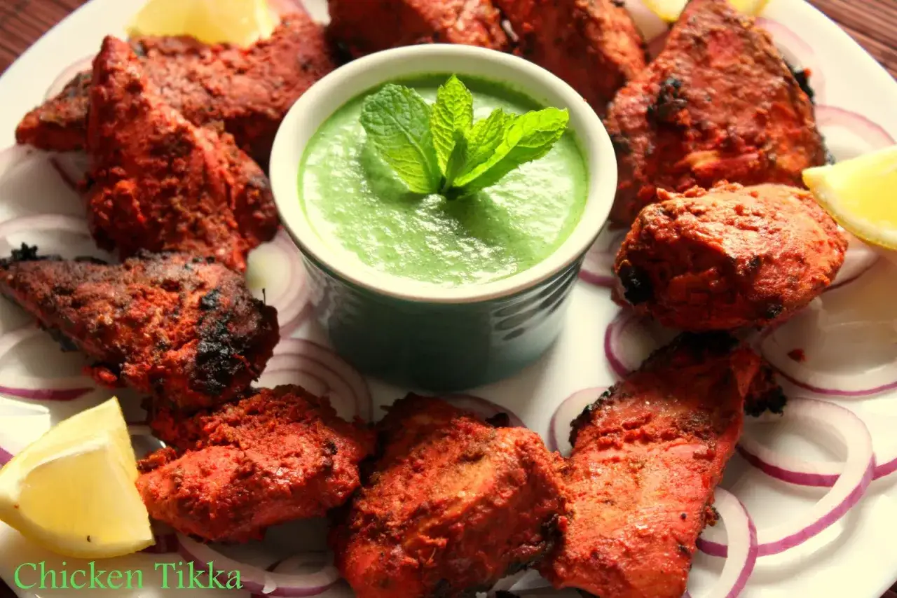 Morceaux de poulet tikka rouges et &eacute;pic&eacute;s, servis avec une sauce verte &agrave; la menthe, des rondelles d'oignon rouge et des quartiers de citron.
