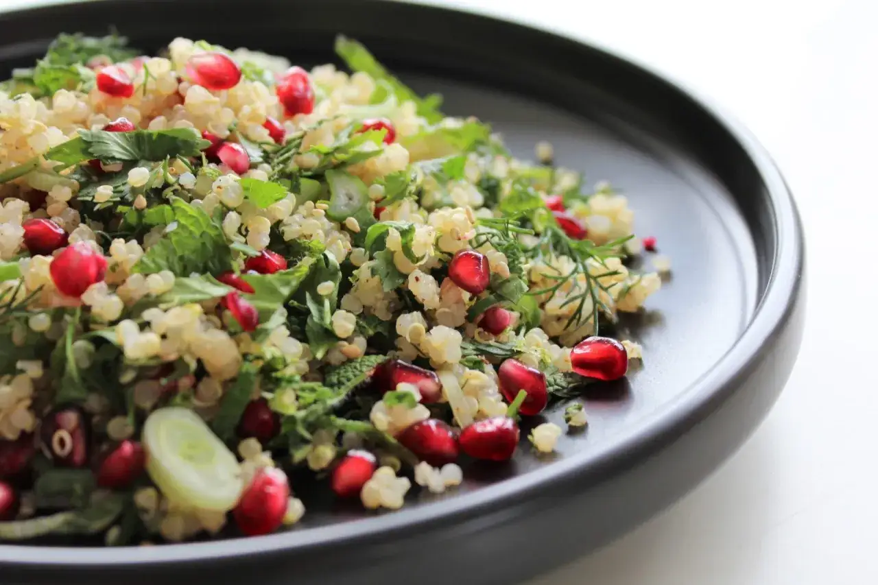 Salade quinoa color&eacute;e avec des graines de grenade, des herbes fra&icirc;ches et des oignons verts, servie dans une assiette noire.
