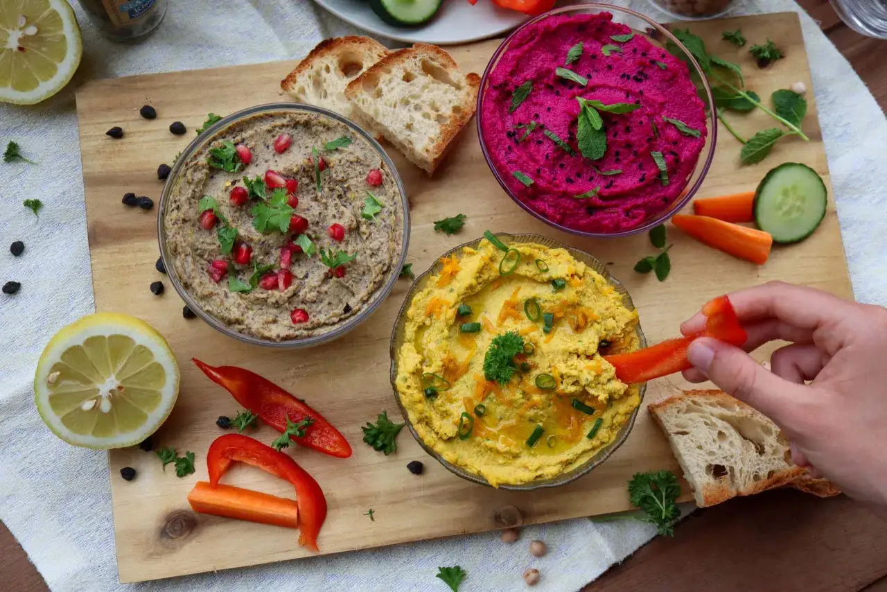 Trois bols de houmous colorés (rose, jaune, brun) avec des garnitures, accompagnés de pain, légumes et citron. Parfait pour découvrir 7 recettes de houmous maison.