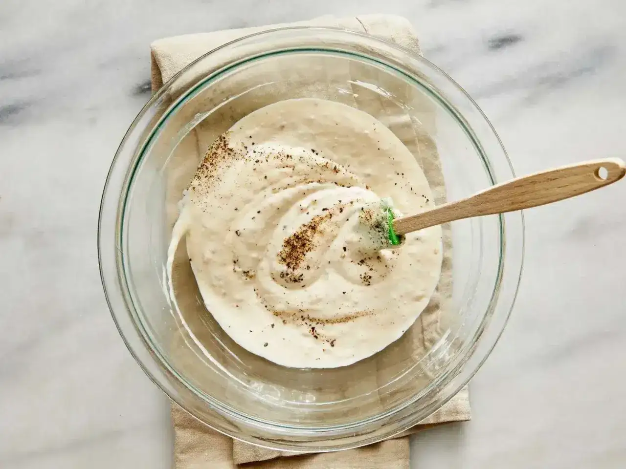 Une spatule en bois m&eacute;lange une sauce c&eacute;sar sans oeuf cr&eacute;meuse et poivr&eacute;e dans un bol en verre.