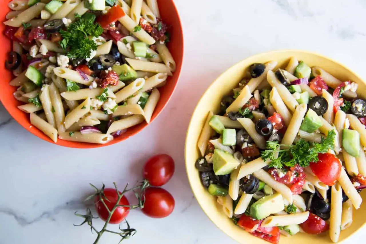 Deux bols color&eacute;s d&eacute;bordent d'une savoureuse salade de p&acirc;tes m&eacute;diterran&eacute;enne, garnie d'avocat, de tomates, d'olives et de feta.