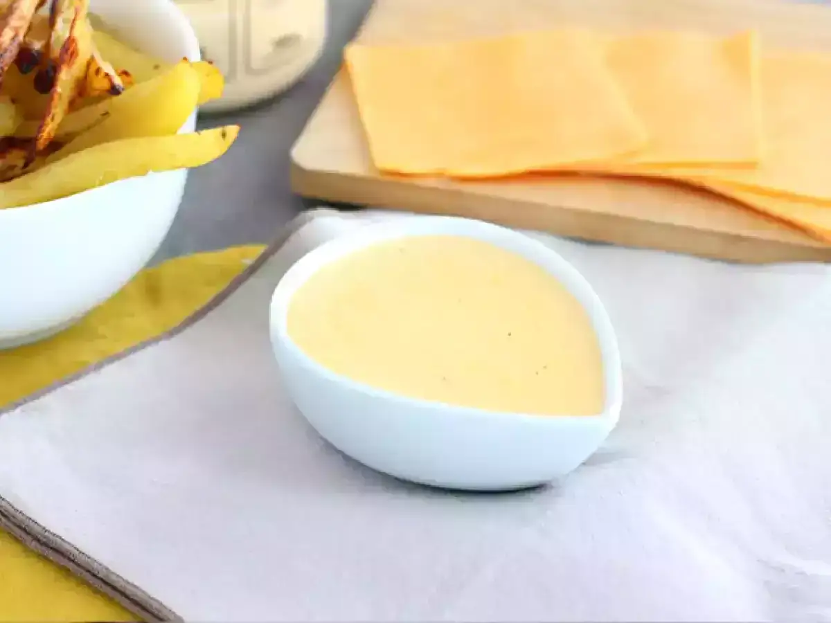 Un bol de sauce fromag&egrave;re cr&eacute;meuse, pr&ecirc;t &agrave; accompagner des frites dor&eacute;es. Des tranches de fromage orange sont dispos&eacute;es sur une planche &agrave; d&eacute;couper en bois.